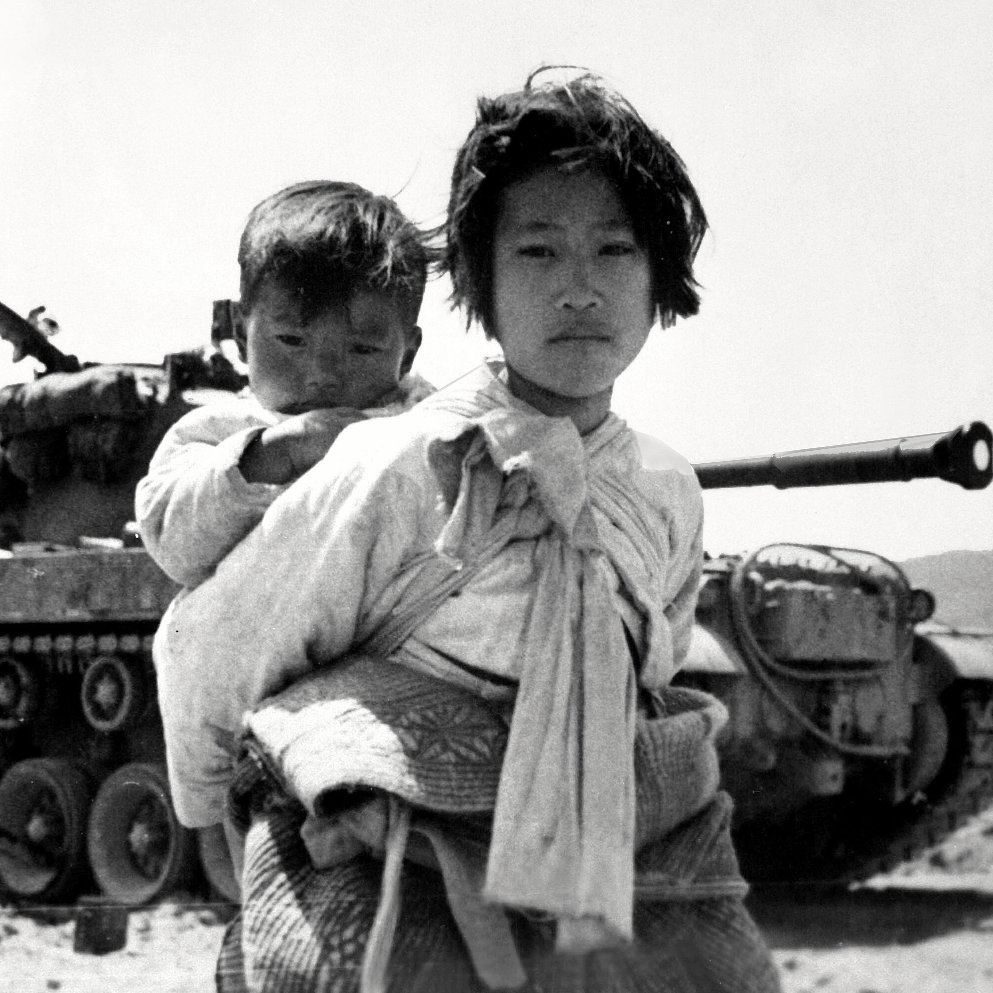 En asiatisk kvinna med ett litet barn på ryggen. De är flyktingar. I bakgrunden står en stridsvagn.