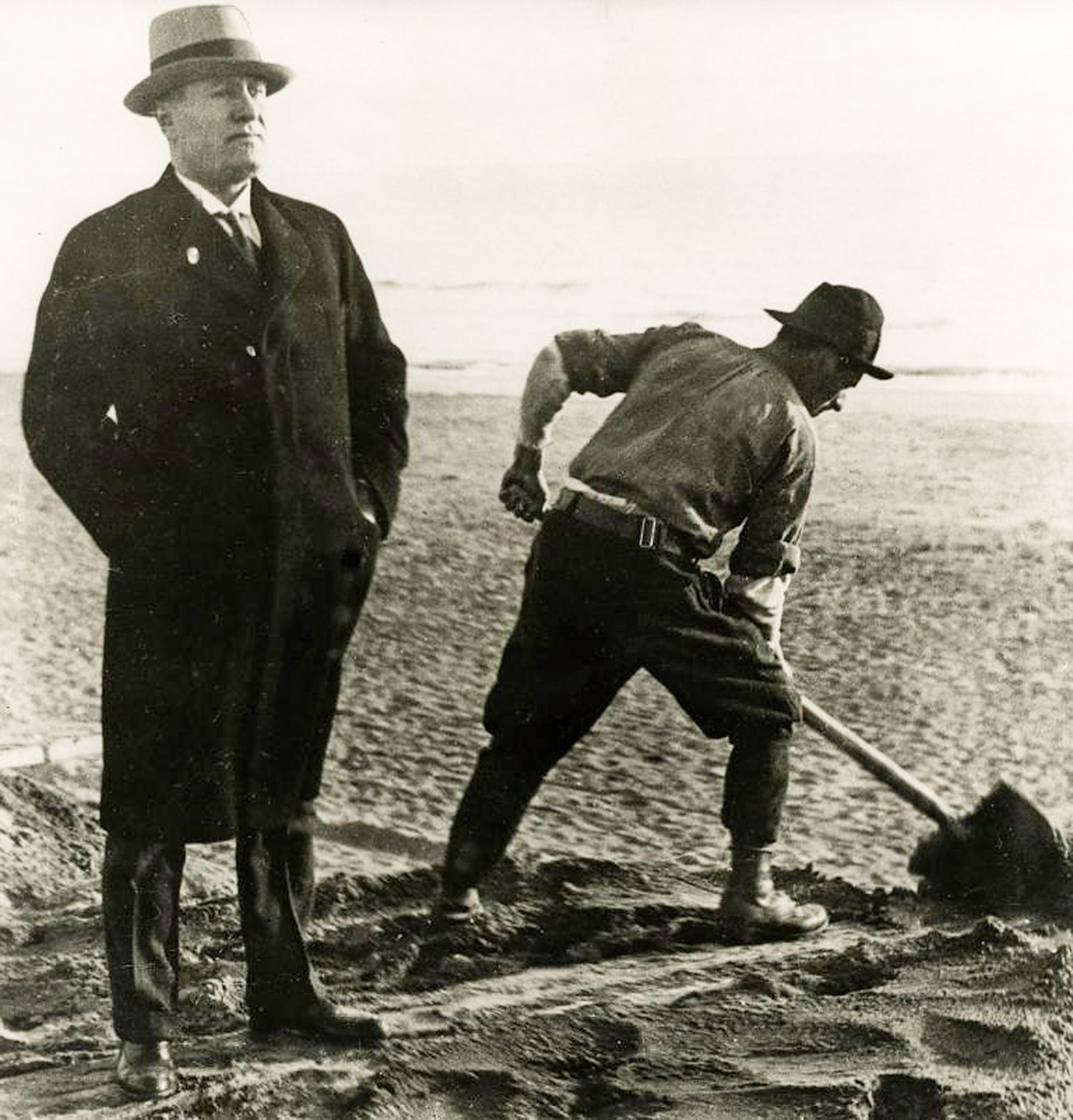En välklädd man i rock och hatt som står bredvid en arbetare som gräver med en spade.