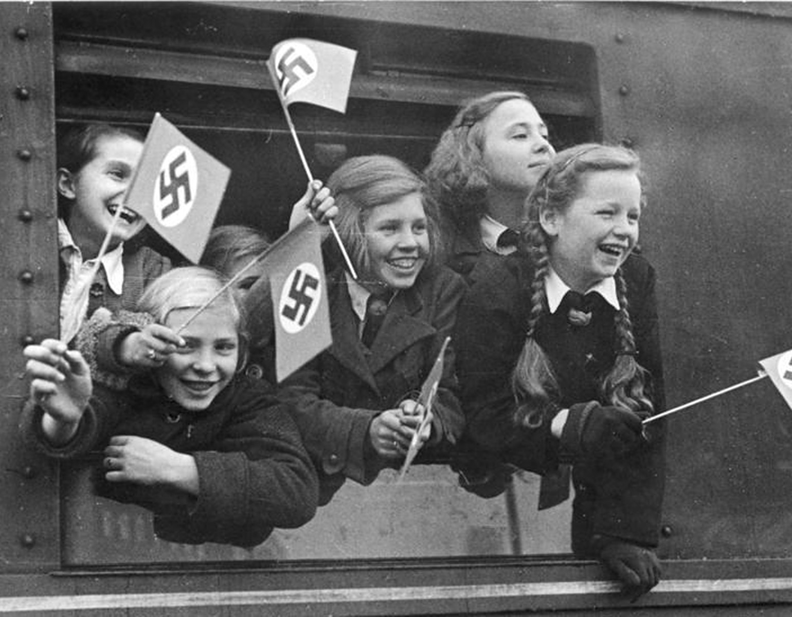 Glada flickor som viftar med små naziflaggor i ett tågfönster.