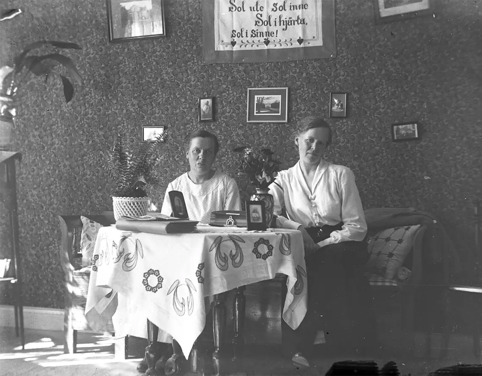 Tv¨å kvinnor som sitter i en soffa i ett vardagsrum. Det är en bonad på väggen och flera fotografier.