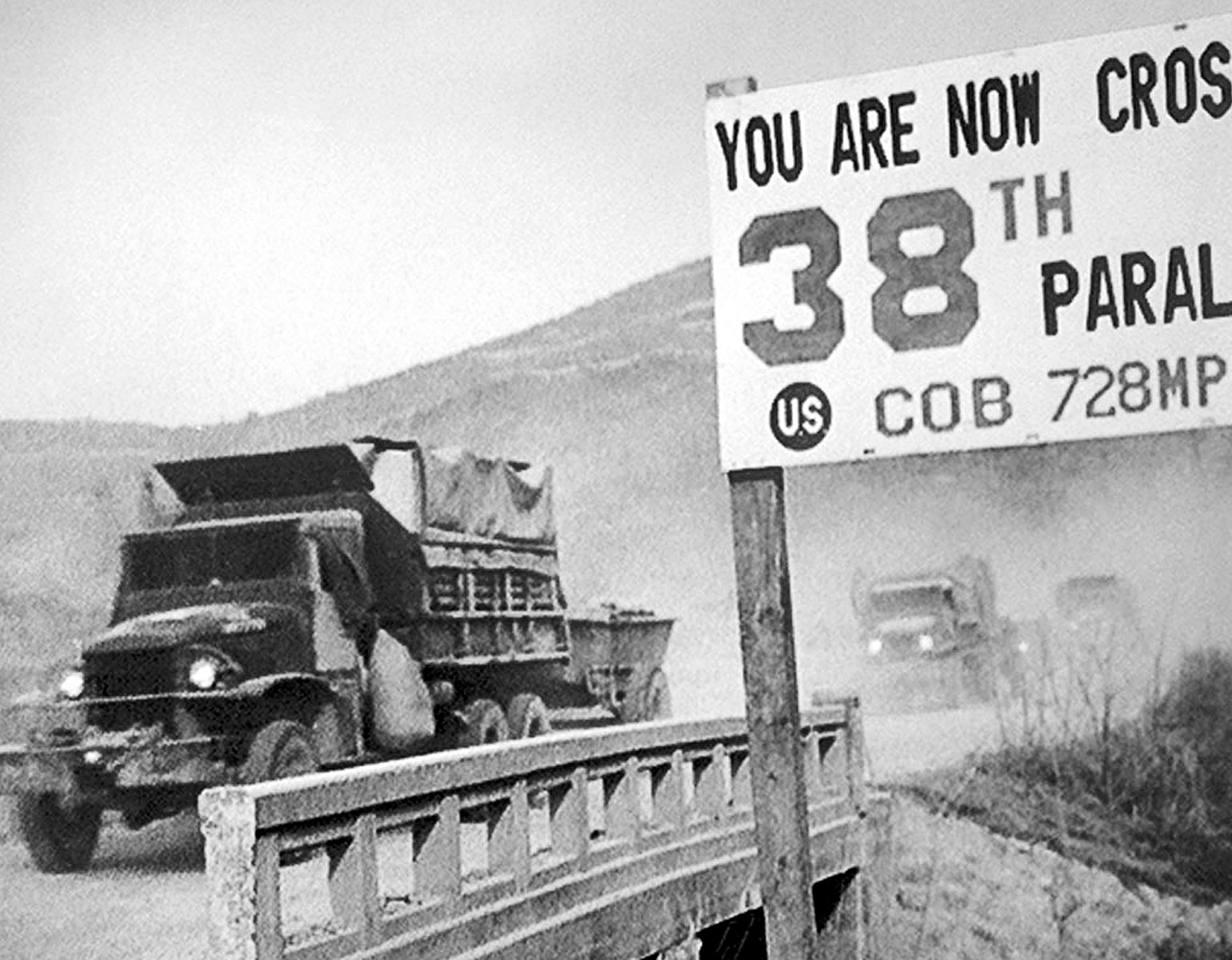 Militärlastbilar som åker över en bro med en skylt som påpekar att de passerar 38e breddgraden.